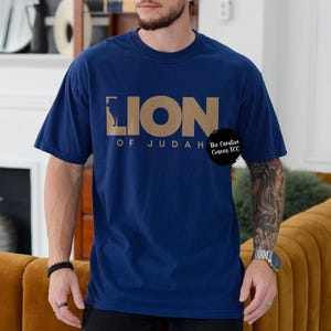K&ouml;nnte beinhalten: Ein marineblaues T-Shirt mit einer goldenen Grafik eines L&ouml;wen und dem Text "Lion of Judah" mit einem kleinen Logo, das "The Creative Canvas TCC" sagt.