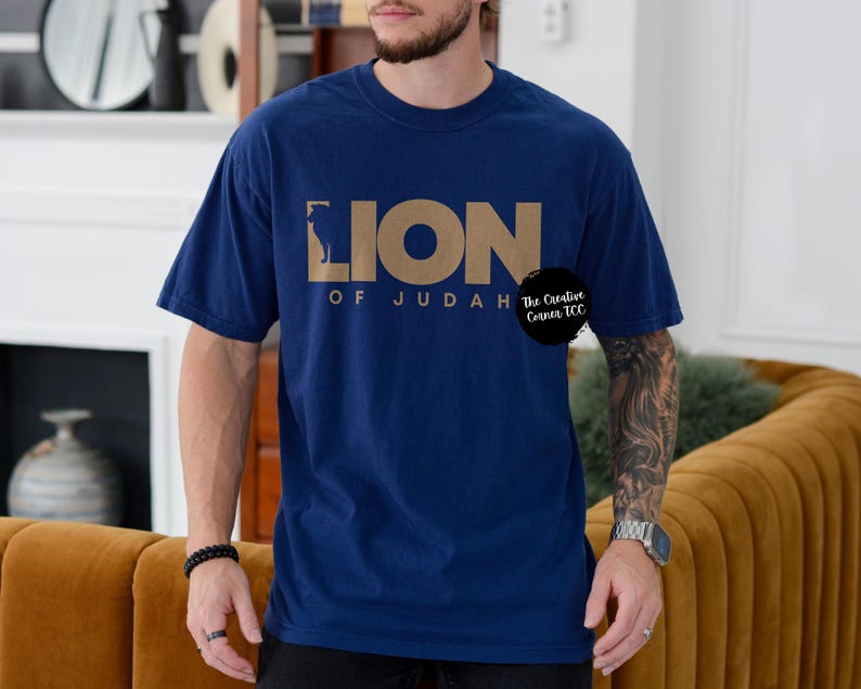 K&ouml;nnte beinhalten: Ein marineblaues T-Shirt mit einer goldenen Grafik eines L&ouml;wen und dem Text "Lion of Judah" mit einem kleinen Logo, das "The Creative Canvas TCC" sagt.