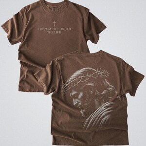 Puede incluir: Camiseta marr&oacute;n con el texto "THE WAY THE TRUTH THE LIFE" y un s&iacute;mbolo de cruz. La parte trasera de la camiseta presenta una ilustraci&oacute;n detallada en blanco y negro de una persona con una corona de espinas. La camiseta tiene un aspecto vintage y desgastado.