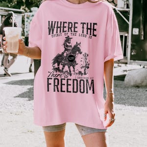 May include: A light pink t-shirt with the text "WHERE THE SPIRIT OF THE LORD IS" above a graphic of a person riding a horse. Below the graphic, the text reads "THERE'S FREEDOM" and "2 CORINTHIANS 3:17".