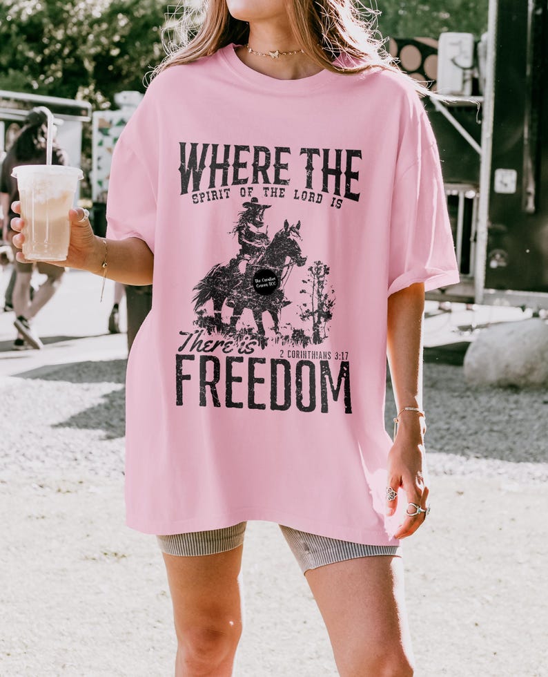 May include: A light pink t-shirt with the text "WHERE THE SPIRIT OF THE LORD IS" above a graphic of a person riding a horse. Below the graphic, the text reads "THERE'S FREEDOM" and "2 CORINTHIANS 3:17".