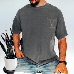 May include: Dark gray t-shirt featuring a gold-colored bull skull graphic on the upper chest. The text "PSALM 90" is printed below the skull. The shirt has a relaxed fit and short sleeves.