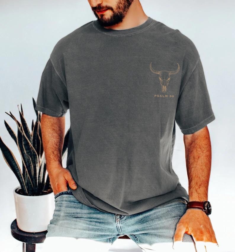 May include: Dark gray t-shirt featuring a gold-colored bull skull graphic on the upper chest. The text "PSALM 90" is printed below the skull. The shirt has a relaxed fit and short sleeves.