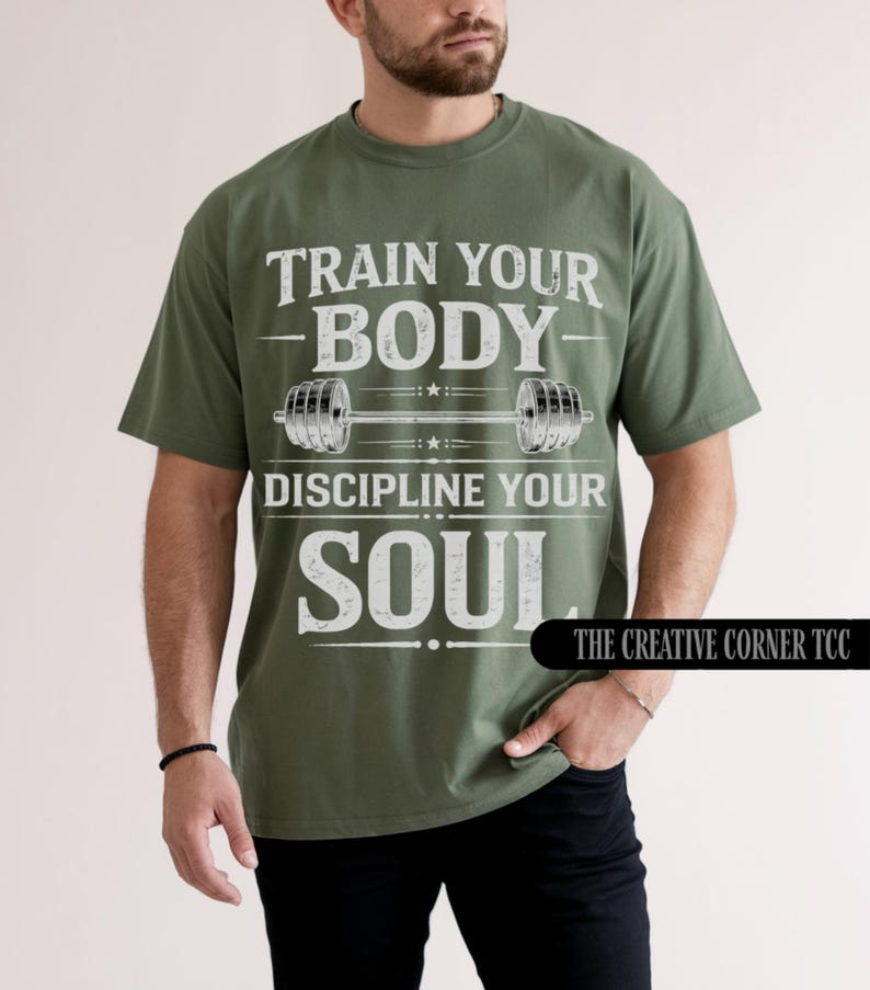 May include: Olive green t-shirt with the text "TRAIN YOUR BODY, DISCIPLINE YOUR SOUL" in white. A barbell graphic is centered between the phrases. The Creative Corner TCC logo is in the bottom right corner.