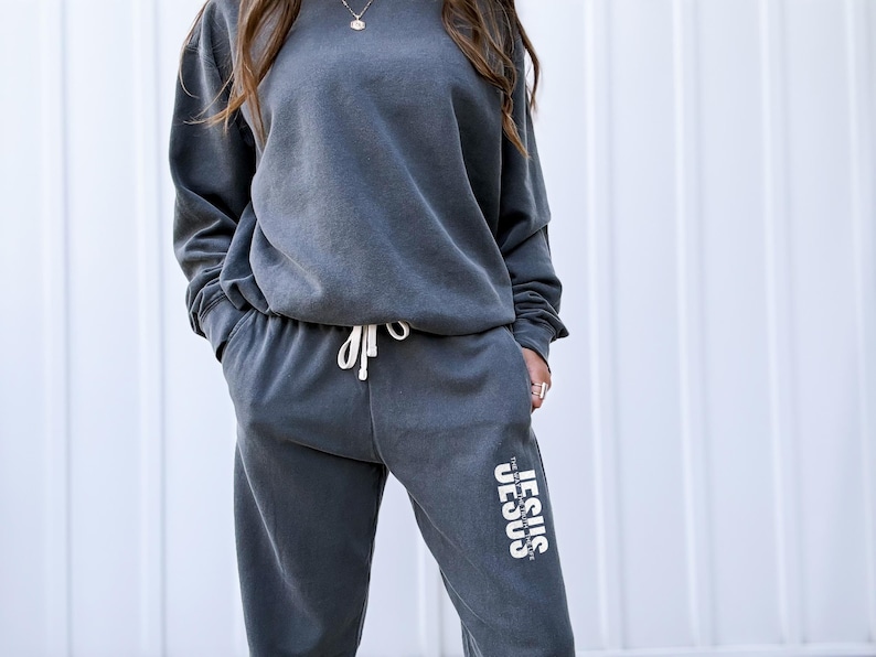 May include: Charcoal gray sweatshirt and sweatpants set. The long-sleeved top has a crew neck and the pants have a drawstring waist. The text "The Jesus" is printed vertically on the left pant leg in white.