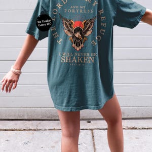 May include: A teal blue t-shirt with a graphic of an eagle with wings spread. The text "The Lord is my refuge and my fortress" is printed in a circle around the eagle. The text "I will never be shaken Psalm 91" is printed below the eagle.