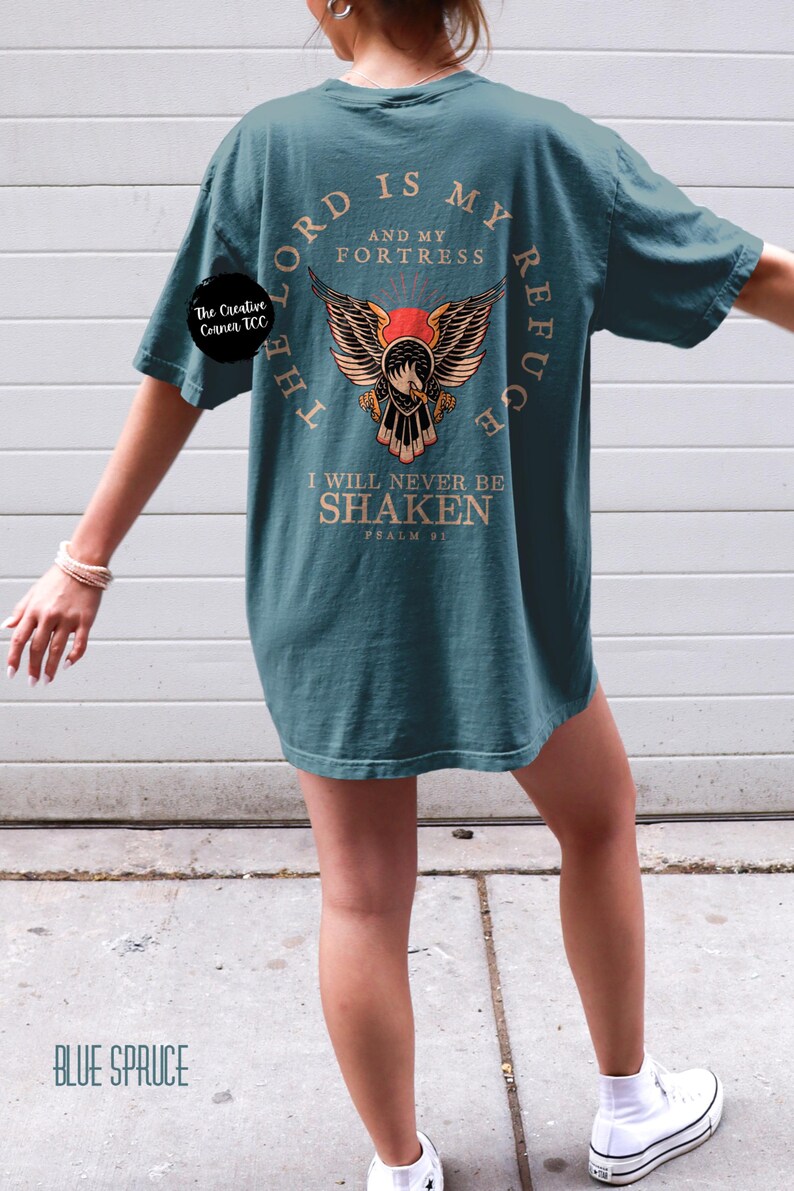 May include: A teal blue t-shirt with a graphic of an eagle with wings spread. The text "The Lord is my refuge and my fortress" is printed in a circle around the eagle. The text "I will never be shaken Psalm 91" is printed below the eagle.