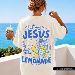 May include: An off-white short-sleeve t-shirt featuring a blue and yellow graphic on the back. The text reads, but my JESUS he be makin' LEMONADE, accompanied by illustrations of a mason jar filled with lemonade, ice cubes, and lemons.