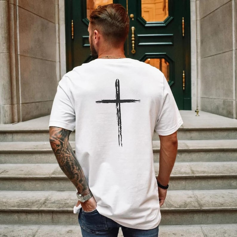 May include: White short-sleeve t-shirt with a black cross design on the back. The cross is a simple, brushstroke style. The shirt is a classic crew neck style.