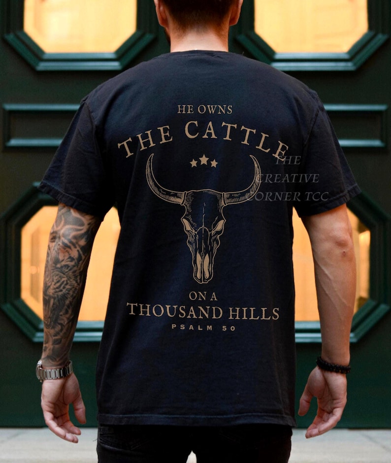 May include: Black t-shirt with a gold graphic of a bull skull and the text "He owns the cattle on a thousand hills Psalm 50". The shirt is a crew neck and has short sleeves.