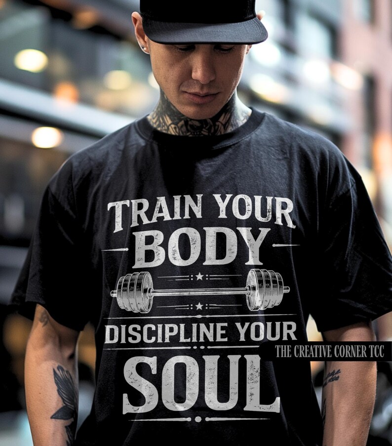 Op de afbeelding: Zwart T-shirt met de witte tekst "TRAIN YOUR BODY - DISCIPLINE YOUR SOUL". Een halterafbeelding is gecentreerd onder "BODY". The Creative Corner TCC staat rechtsonder afgedrukt. De persoon draagt een zwarte pet.