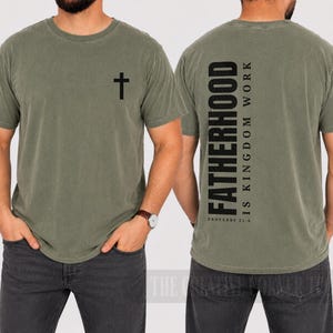 May include: Olive green t-shirt with a black cross on the front. The back of the shirt features the word "FATHERHOOD" in large, vertical black letters, with "IS KINGDOM WORK" and "PROVERBS 22:6" printed below.