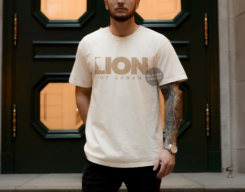 K&ouml;nnte beinhalten: Ein beiges T-Shirt mit einer braunen Grafik eines L&ouml;wen und dem Text "Lion of Judah" und "The Greatest Commandment CCG".