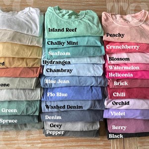 Op de afbeelding: Een stapel opgevouwen t-shirts in verschillende kleuren, waaronder wit, ivoor en perzik. Elk shirt is gelabeld met zijn kleurnaam, zoals "Island Reef", "Chalky Mint" en "Berry". De shirts zijn netjes gerangschikt en tonen een reeks tinten.