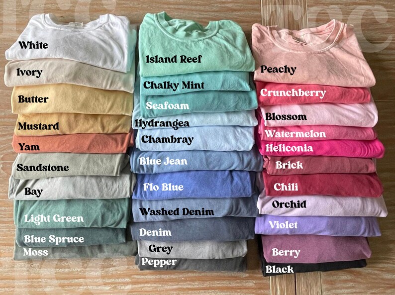 Op de afbeelding: Een stapel opgevouwen t-shirts in verschillende kleuren, waaronder wit, ivoor en perzik. Elk shirt is gelabeld met zijn kleurnaam, zoals "Island Reef", "Chalky Mint" en "Berry". De shirts zijn netjes gerangschikt en tonen een reeks tinten.