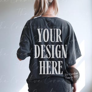 May include: A black oversized t-shirt with the text "YOUR DESIGN HERE" printed on the back. The shirt is being worn by a person with their back to the camera.