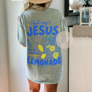 May include: A sage green short-sleeve t-shirt featuring a back graphic with blue and yellow text that reads but my JESUS he be makin LEMONADE. The design includes illustrations of a mason jar filled with lemonade, lemon slices, and small stars.