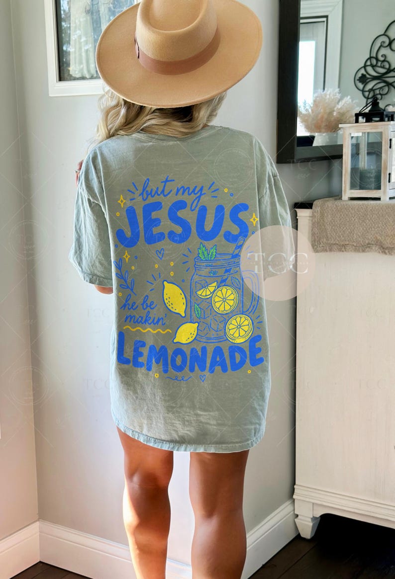 May include: A sage green short-sleeve t-shirt featuring a back graphic with blue and yellow text that reads but my JESUS he be makin LEMONADE. The design includes illustrations of a mason jar filled with lemonade, lemon slices, and small stars.