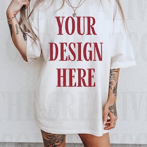 Puede incluir: Camiseta de gran tamaño de color crema con el texto "YOUR DESIGN HERE" en negrita, burdeos. La camiseta tiene mangas cortas y un ajuste relajado. La modelo lleva botas de vaquero rojas.