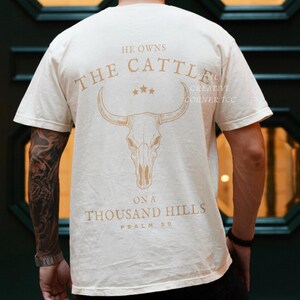 May include: Cream-colored t-shirt with a graphic of a bull skull and the text "He Owns The Cattle On A Thousand Hills Psalm 50" in a light brown color. The shirt is short-sleeved.
