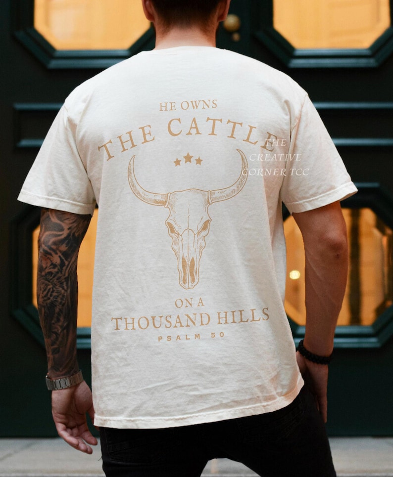 May include: Cream-colored t-shirt with a graphic of a bull skull and the text "He Owns The Cattle On A Thousand Hills Psalm 50" in a light brown color. The shirt is short-sleeved.