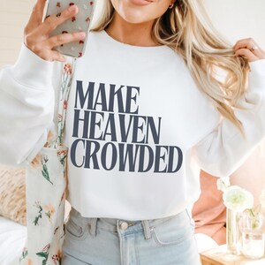 May include: White sweatshirt with the navy blue text "MAKE HEAVEN CROWDED". The sweatshirt is worn with light wash jeans. The image shows a person holding a phone and a floral tote bag.