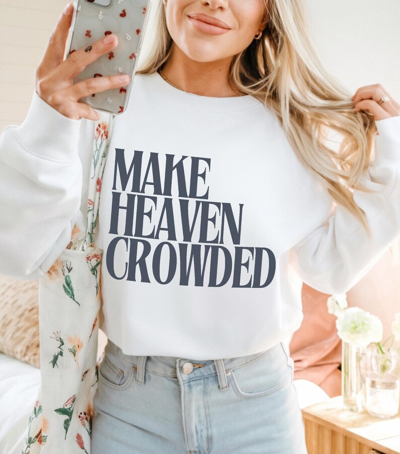 May include: White sweatshirt with the navy blue text "MAKE HEAVEN CROWDED". The sweatshirt is worn with light wash jeans. The image shows a person holding a phone and a floral tote bag.