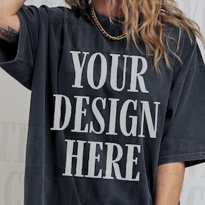 May include: Charcoal grey t-shirt featuring the text "YOUR DESIGN HERE" in bold, white lettering. The shirt has short sleeves and a vintage, washed-out look. The style is casual and relaxed.