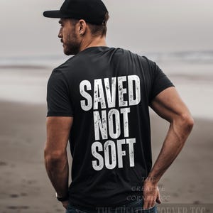 May include: Black t-shirt with the white text "SAVED NOT SOFT" on the back. The shirt is worn with a black baseball cap and blue jeans. The Creative Corner TCC is printed at the bottom of the shirt.