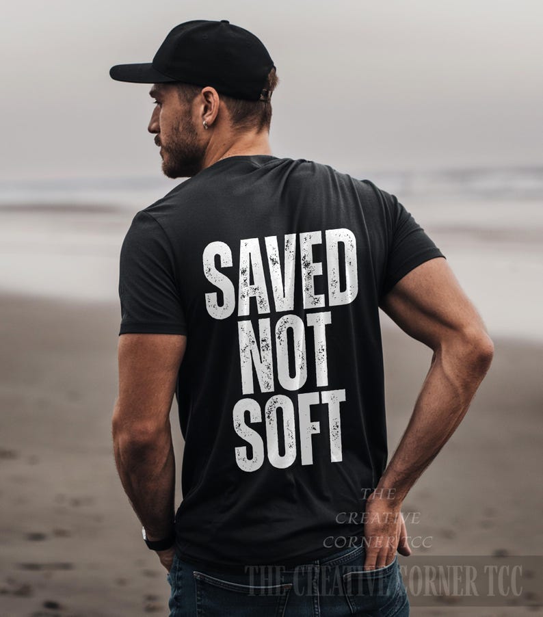 May include: Black t-shirt with the white text "SAVED NOT SOFT" on the back. The shirt is worn with a black baseball cap and blue jeans. The Creative Corner TCC is printed at the bottom of the shirt.