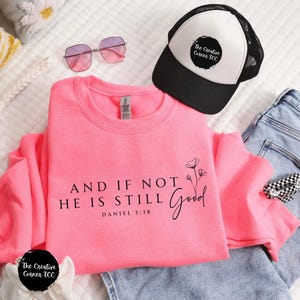 May include: A bright pink sweatshirt with the text "AND IF NOT HE IS STILL GOOD" and "DANIEL 3:18". The image also shows a black and white trucker hat, sunglasses, and a pair of blue jeans.