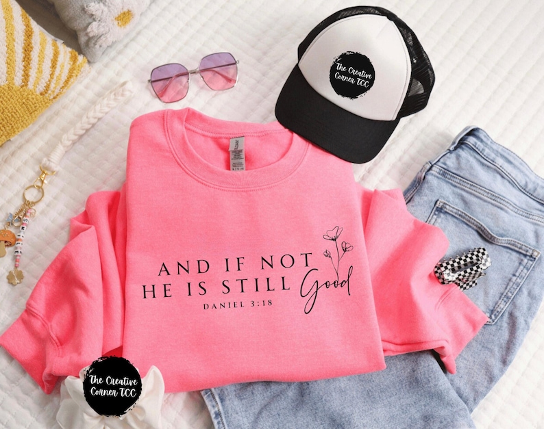 May include: A bright pink sweatshirt with the text "AND IF NOT HE IS STILL GOOD" and "DANIEL 3:18". The image also shows a black and white trucker hat, sunglasses, and a pair of blue jeans.