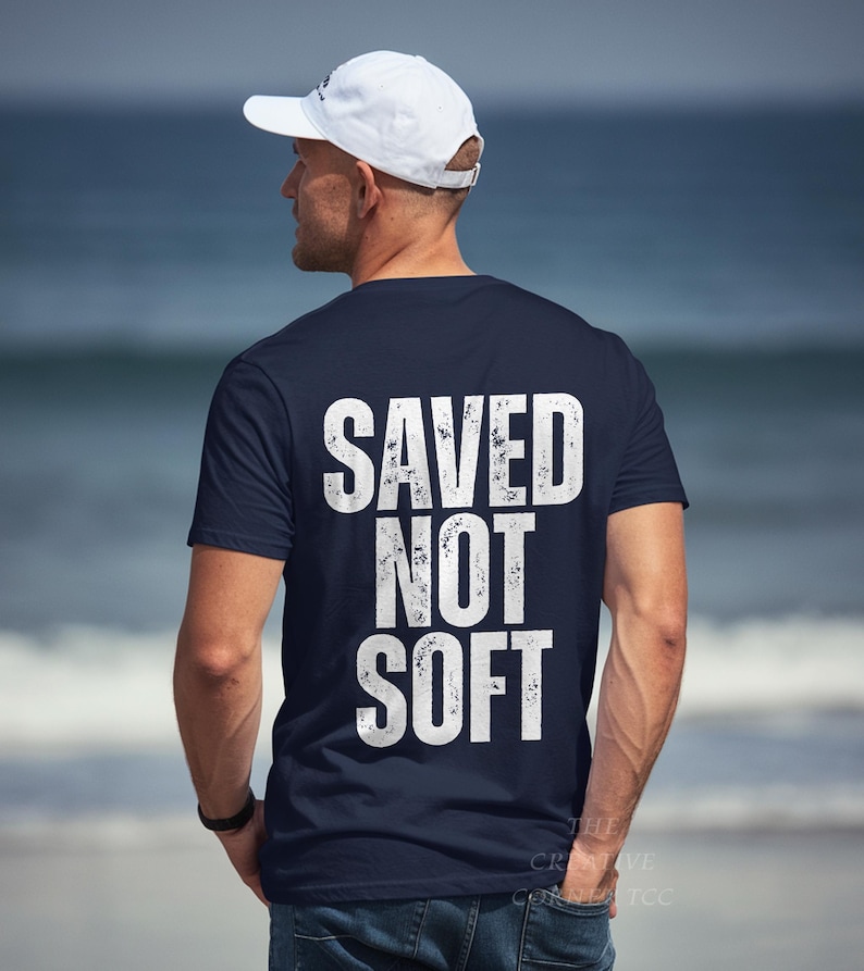 May include: Navy blue t-shirt with the white text "SAVED NOT SOFT" in a distressed font. The shirt is short-sleeved and worn by a person wearing a white baseball cap.