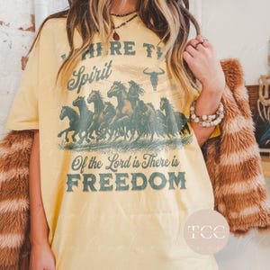 May include: Yellow graphic tee shirt with a vintage design featuring a herd of horses running across a field. The text on the shirt reads "Where the Spirit of the Lord is There is Freedom".