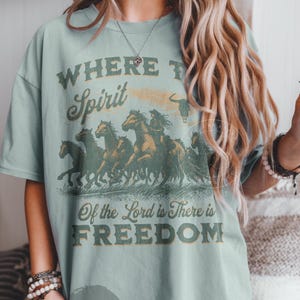 May include: A light blue t-shirt with a vintage graphic design featuring a group of horses running. The text on the shirt reads "Where the Spirit of the Lord is There is Freedom".