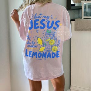 May include: A light pink short-sleeve t-shirt featuring a blue and yellow graphic on the back. The text reads but my JESUS he be makin' LEMONADE, accompanied by illustrations of a mason jar filled with lemonade, ice cubes, and lemon slices.