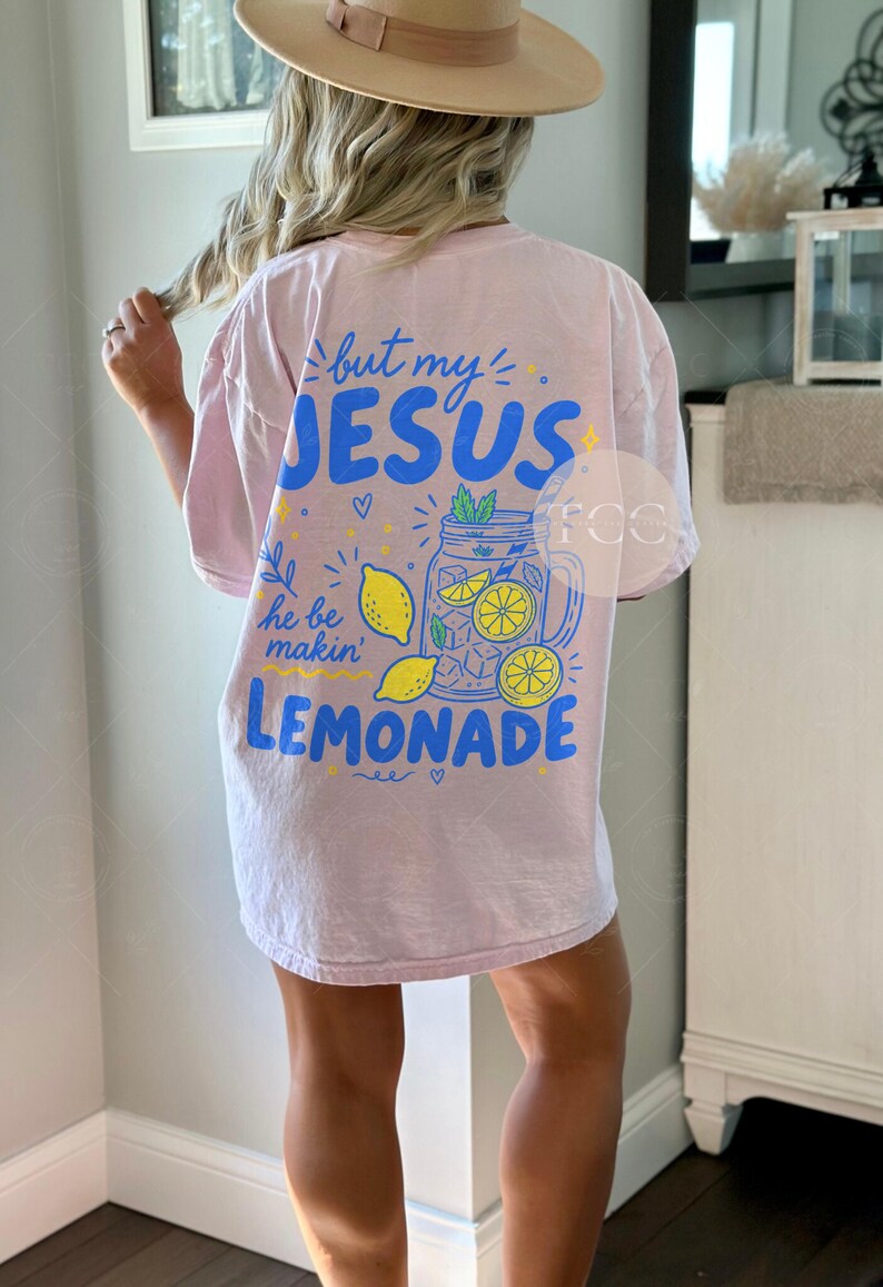 May include: A light pink short-sleeve t-shirt featuring a blue and yellow graphic on the back. The text reads but my JESUS he be makin' LEMONADE, accompanied by illustrations of a mason jar filled with lemonade, ice cubes, and lemon slices.