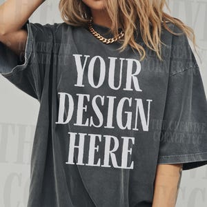 Könnte beinhalten: Dunkelgraues, übergroßes T-Shirt mit dem Text "YOUR DESIGN HERE" in fetten weißen Buchstaben. Das Shirt hat kurze Ärmel und einen Vintage-Look. Eine goldene Halskette ist sichtbar.