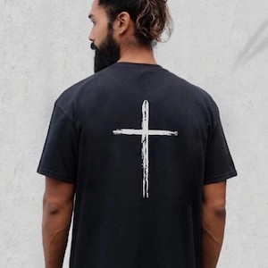 May include: Black t-shirt with a white, brushstroke-style cross design on the back. The shirt is a solid black color and appears to be made of a soft material. The cross is centered on the back of the shirt.
