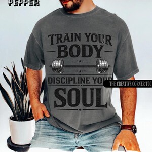Op de afbeelding: Donkergrijs t-shirt met de tekst "TRAIN YOUR BODY, DISCIPLINE YOUR SOUL" en een halterafbeelding. Het shirt heeft een vintage uitstraling. De merknamen "PEPPER" en "THE CREATIVE CORNER TCC" staan linksboven en rechtsonder.