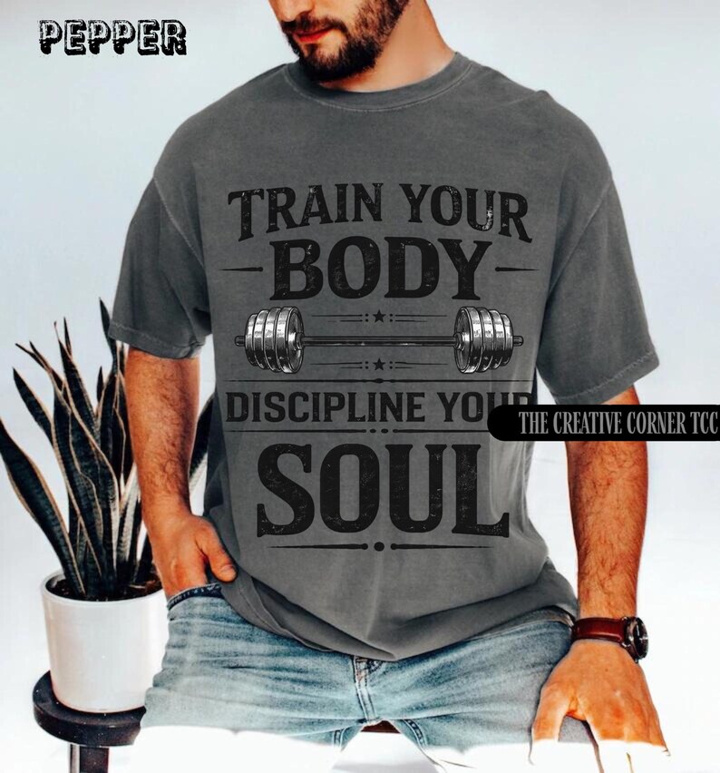 Op de afbeelding: Donkergrijs t-shirt met de tekst "TRAIN YOUR BODY, DISCIPLINE YOUR SOUL" en een halterafbeelding. Het shirt heeft een vintage uitstraling. De merknamen "PEPPER" en "THE CREATIVE CORNER TCC" staan linksboven en rechtsonder.