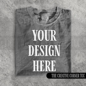 May include: A gray t-shirt with the words "YOUR DESIGN HERE" in large white letters. The shirt is folded neatly and has a tag that reads "COMFORT COLORS". The text "THE CREATIVE CORNER TCC" is at the bottom.