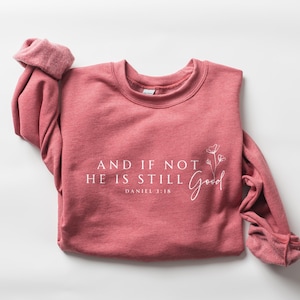 May include: A dusty rose-colored sweatshirt with the text "AND IF NOT HE IS STILL Good" and "DANIEL 3:18" printed in white. A floral design is next to the word "Good". The long sleeves are rolled up. The sweatshirt is made of a soft material.