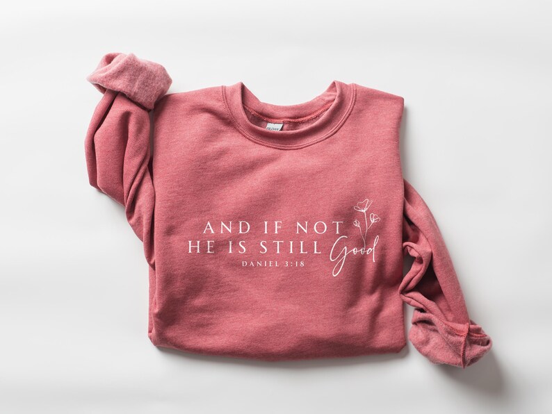 May include: A dusty rose-colored sweatshirt with the text "AND IF NOT HE IS STILL Good" and "DANIEL 3:18" printed in white. A floral design is next to the word "Good". The long sleeves are rolled up. The sweatshirt is made of a soft material.