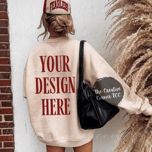 May include: A beige sweatshirt with the text "YOUR DESIGN HERE" printed in red on the back. The model is wearing a red and white baseball cap that says "FEARLESS" in red.