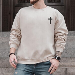 May include: A beige crewneck sweatshirt with a black cross graphic on the upper left chest. The long-sleeved top is made of a soft material and is a neutral color. The sweatshirt is paired with blue jeans.