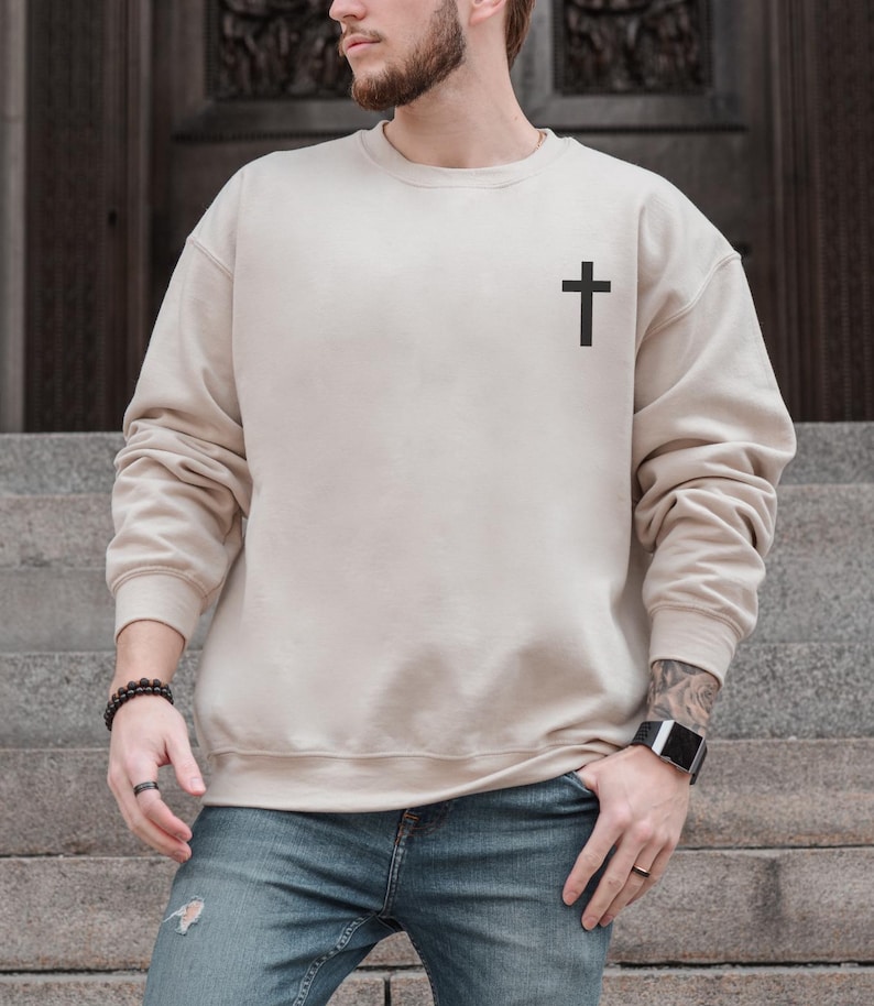 May include: A beige crewneck sweatshirt with a black cross graphic on the upper left chest. The long-sleeved top is made of a soft material and is a neutral color. The sweatshirt is paired with blue jeans.