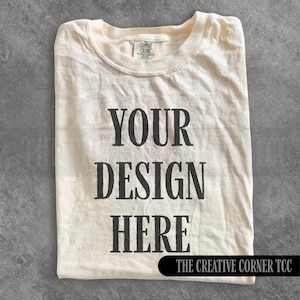 Könnte beinhalten: Cremefarbenes T-Shirt mit dem Text "YOUR DESIGN HERE" in fetter, schwarzer Schrift. Der Text ist mittig auf dem Shirt platziert, mit "THE CREATIVE CORNER TCC" am unteren Rand. Das Shirt ist sauber auf einer grauen Oberfläche gefaltet.