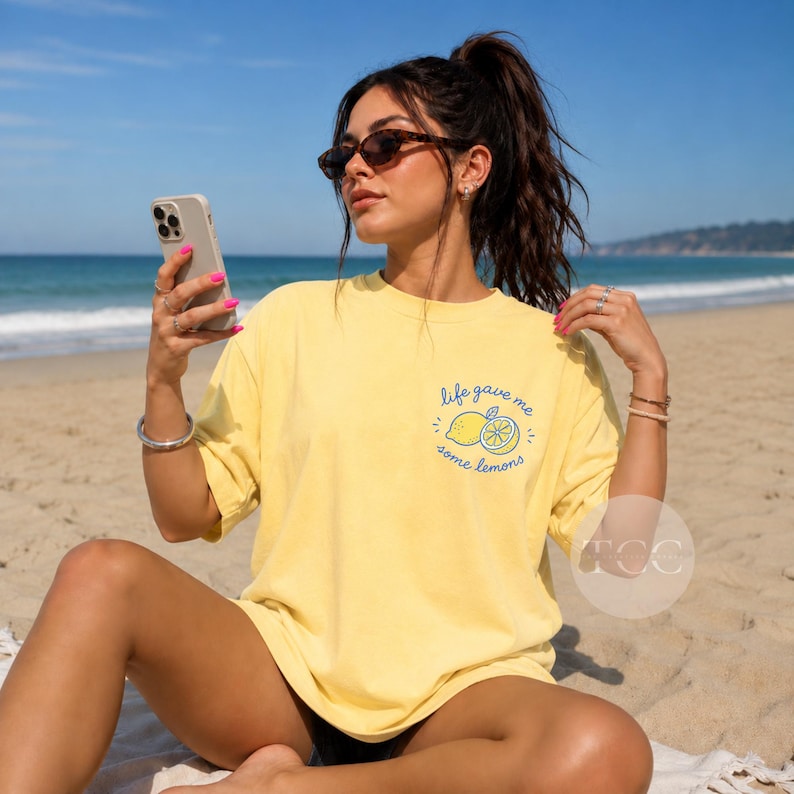 May include: A pale yellow short-sleeve t-shirt featuring a small graphic on the left chest area. The graphic depicts a lemon and a lemon slice with the blue cursive text "life gave me some lemons" arranged in a circular pattern around the fruit.