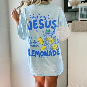 May include: A light blue short-sleeve t-shirt featuring a graphic on the back with the text "but my JESUS he be makin' LEMONADE". The design includes a blue mason jar filled with yellow lemon slices, ice cubes, and green mint leaves.